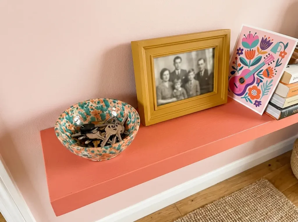 Ceramic key bowl, mustard-framed family photograph, and illustrated postcard on a coral shelf.